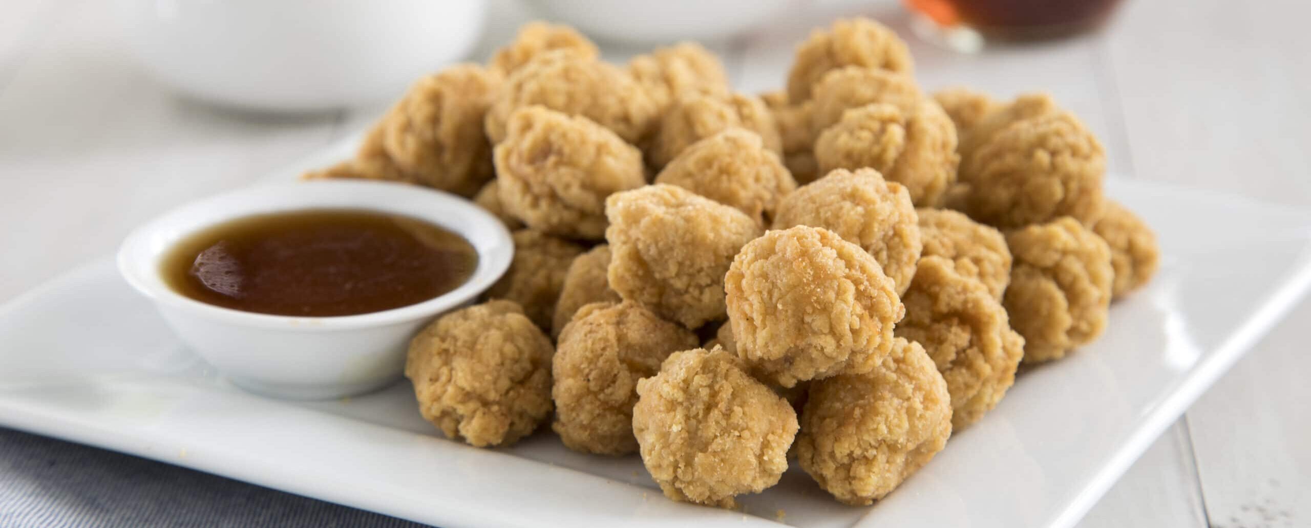 Plate of fried chicken nuggets with a dipping sauce, fruit in the background