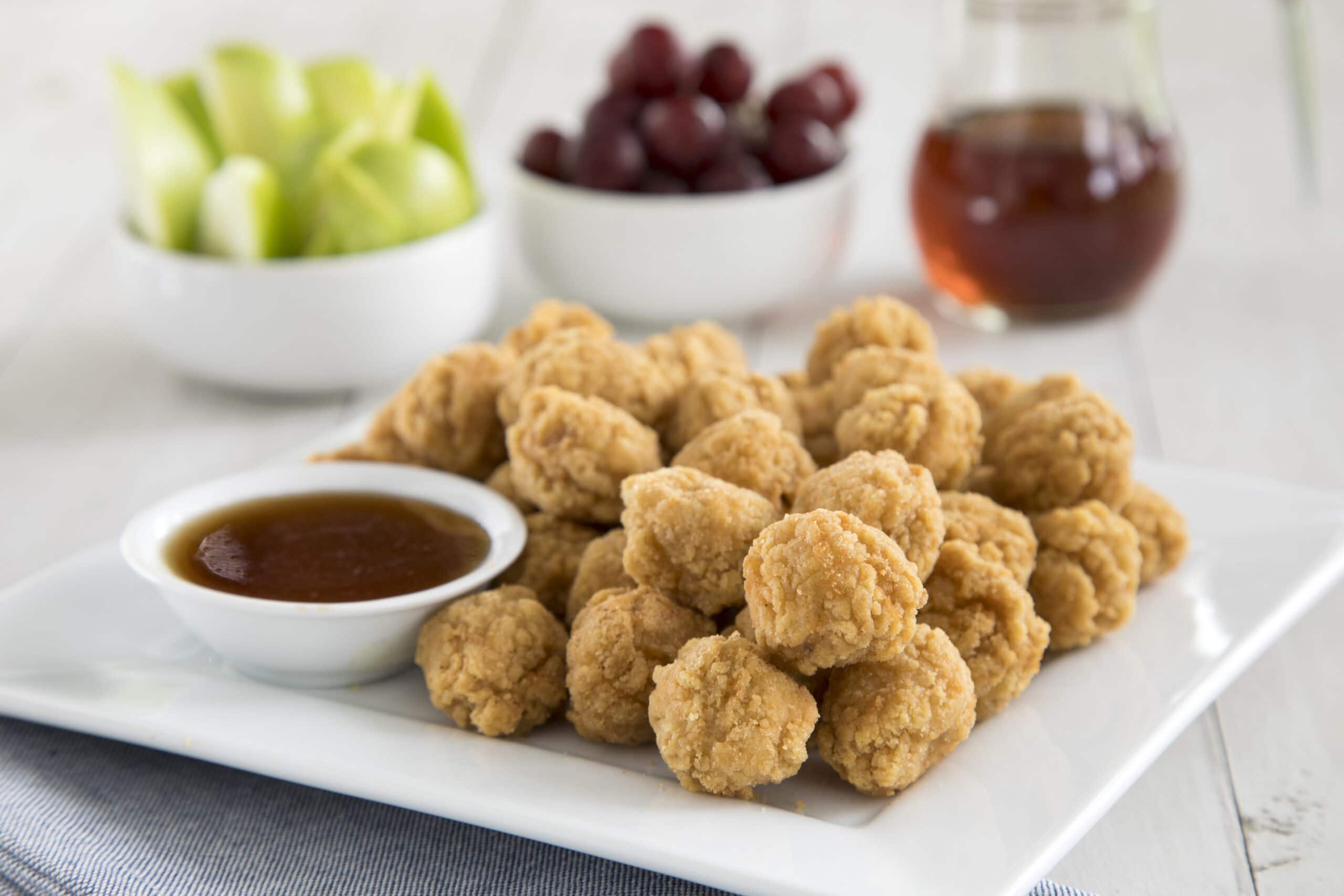 Plate of fried chicken nuggets with a dipping sauce, fruit in the background