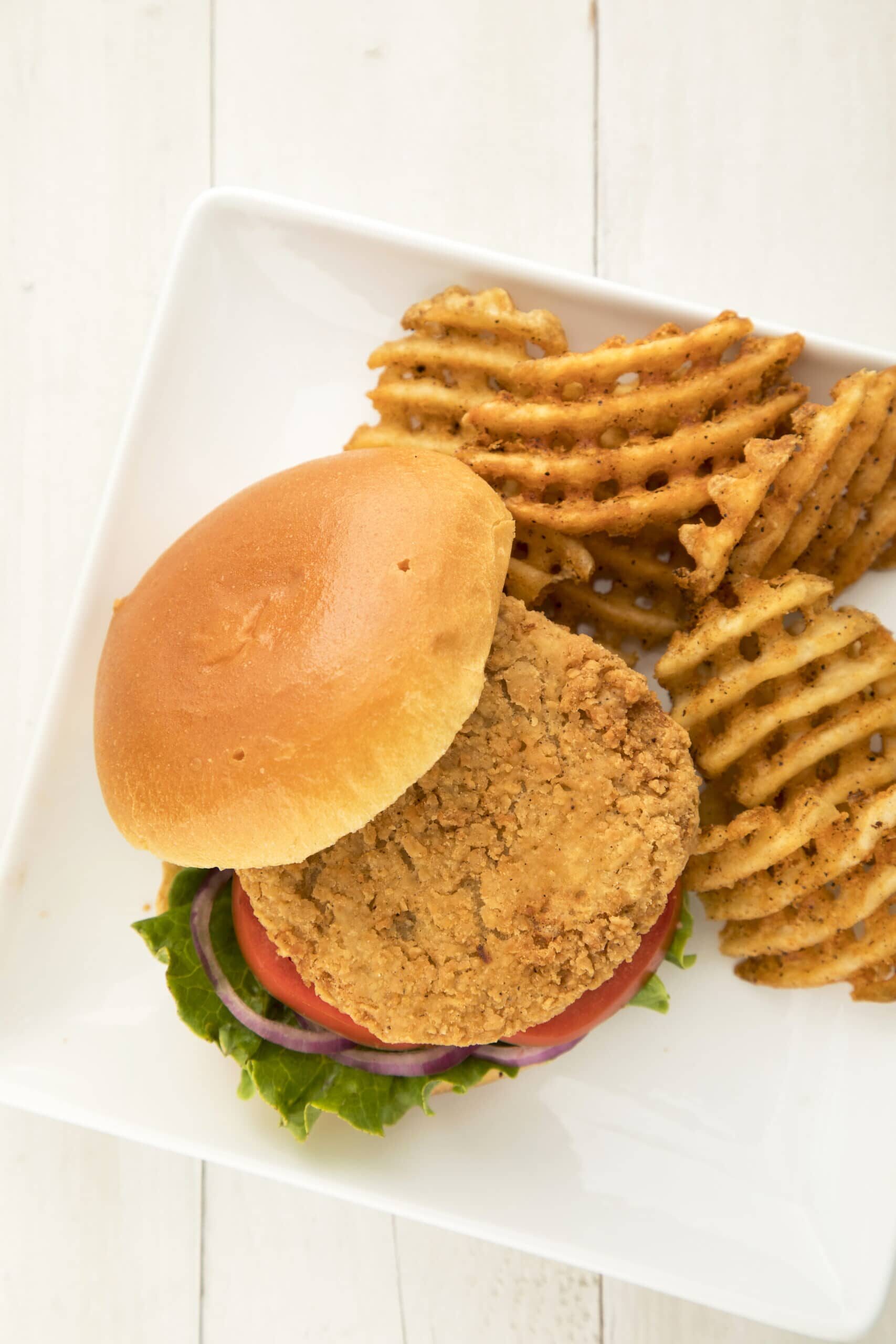 Chicken sandwich on a bun with tomatoes, onions, and lettuce on a plate, with french fries