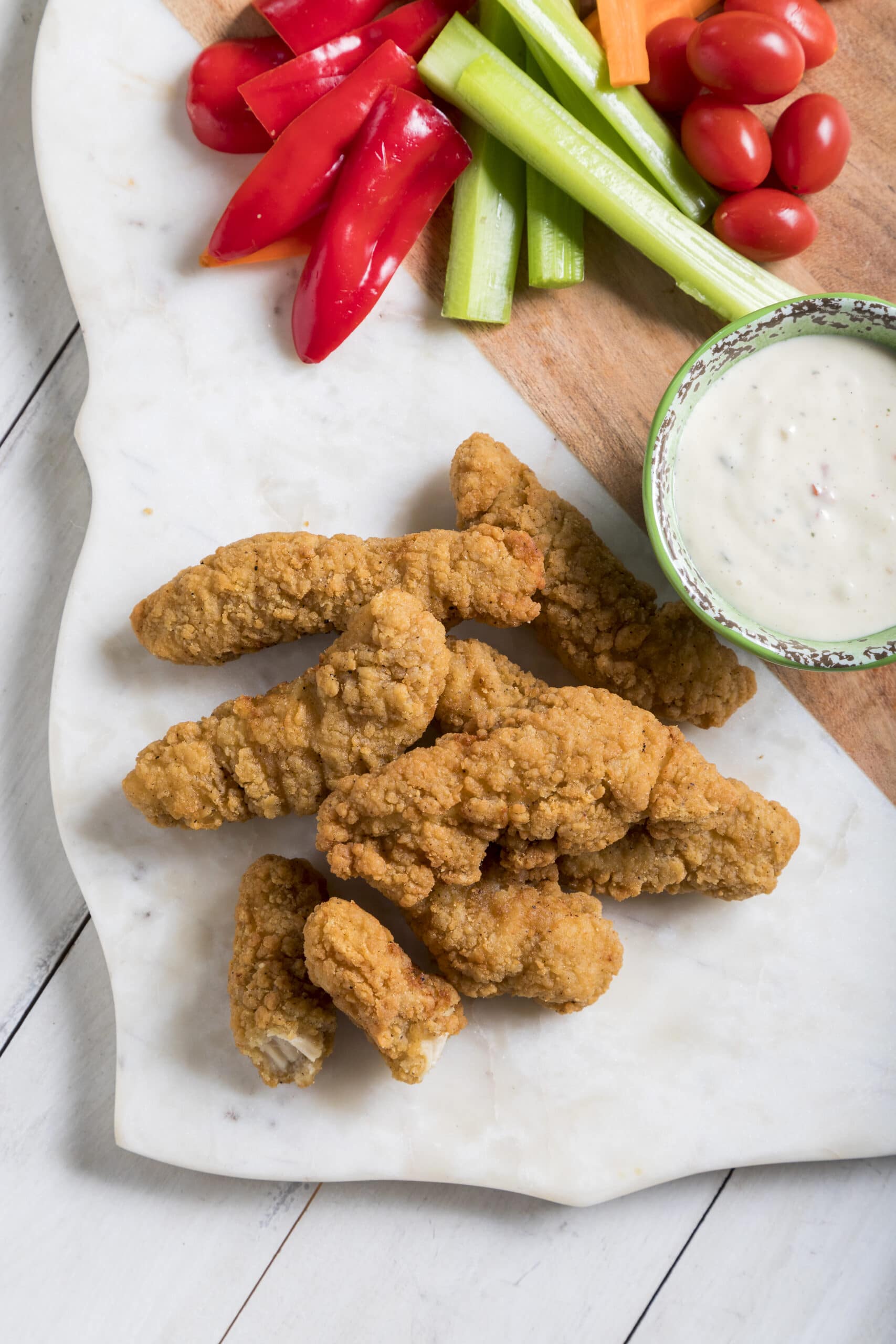 Chicken tenders on a marble serving tray with peppers, celery, tomatoes, and dipping sauce