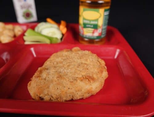 Chicken breast fillet on school lunch tray with raw veggie side and bottle of apple juice